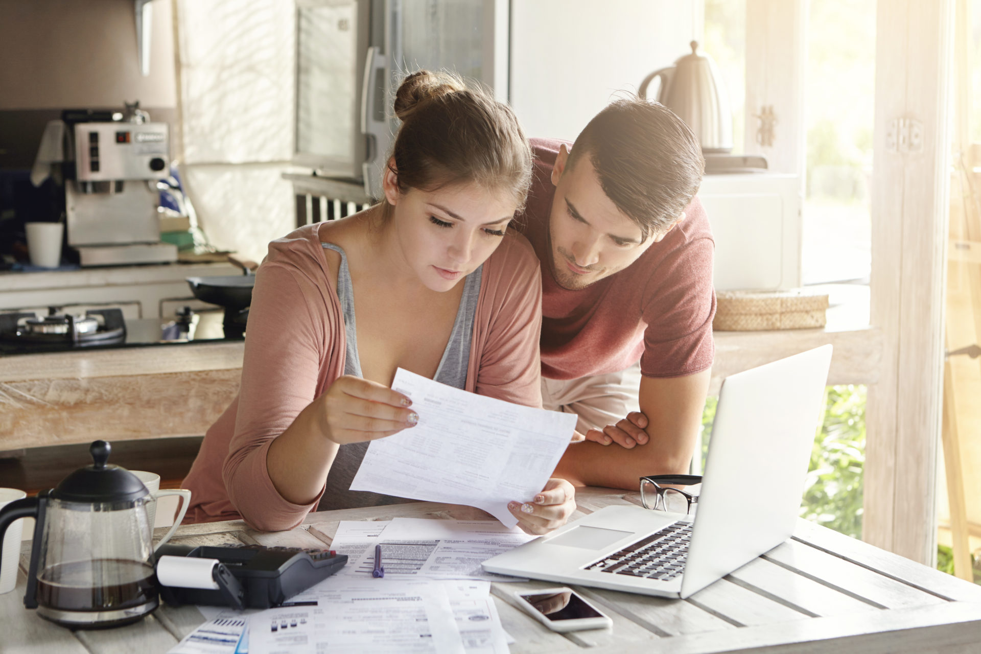 Young couple managing finances, reviewing their bank accounts using laptop computer and calculator at modern kitchen. Woman and man doing paperwork together, paying taxes online on notebook pc